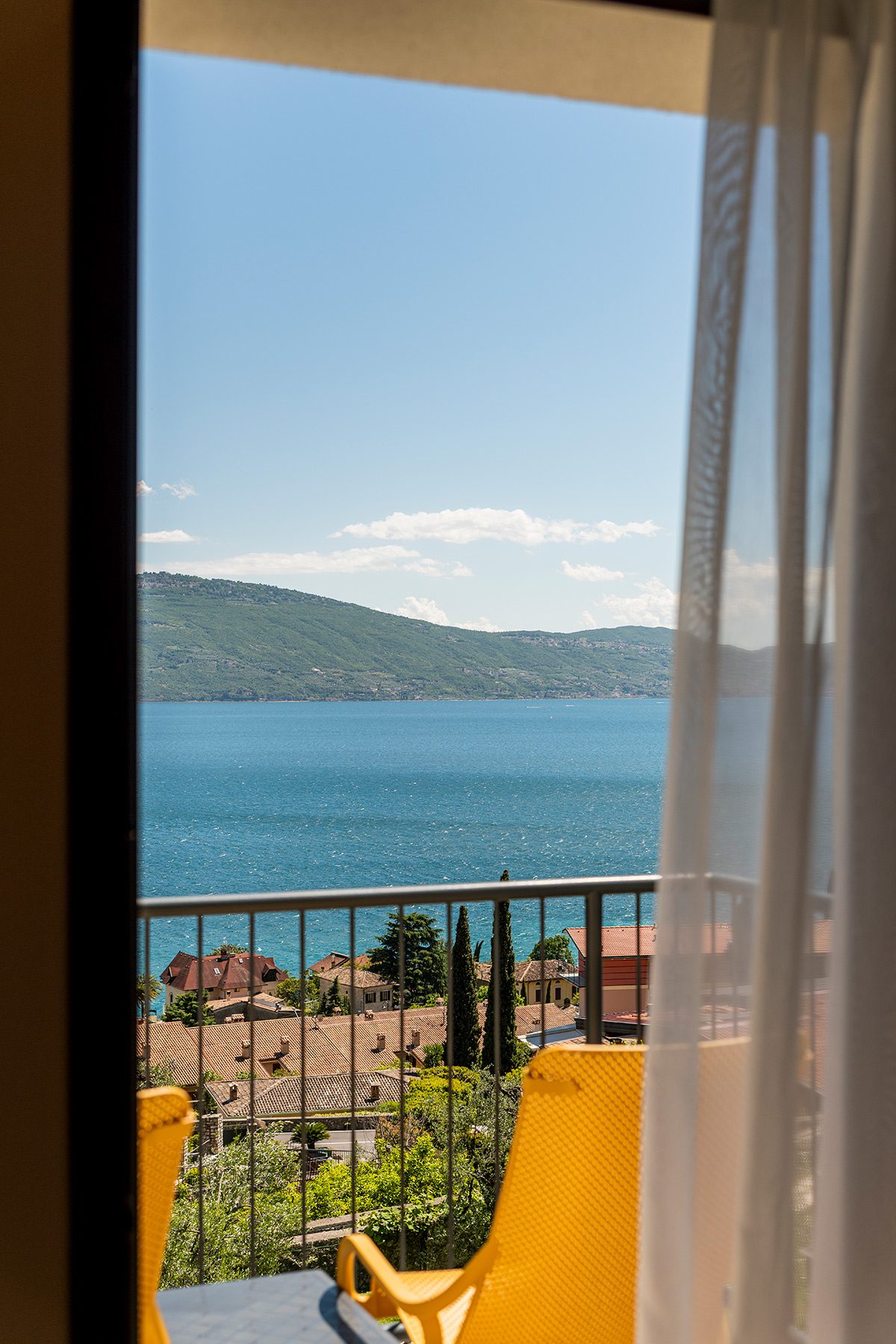 Vista lago da balcone con sedie gialle, tetti e colline al sole