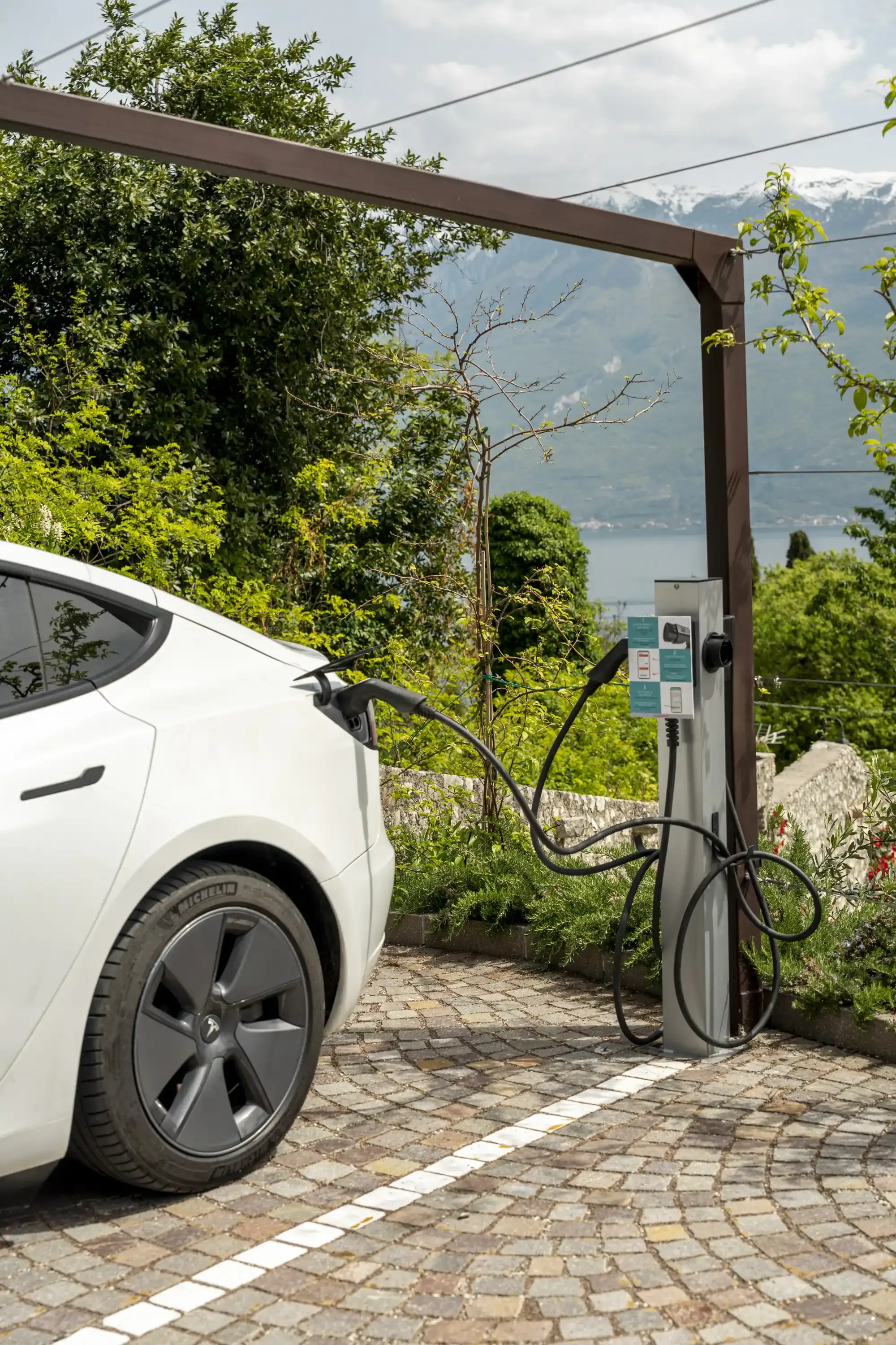 Weißes Elektroauto lädt an Ladestation im Freien mit See und Bergen.