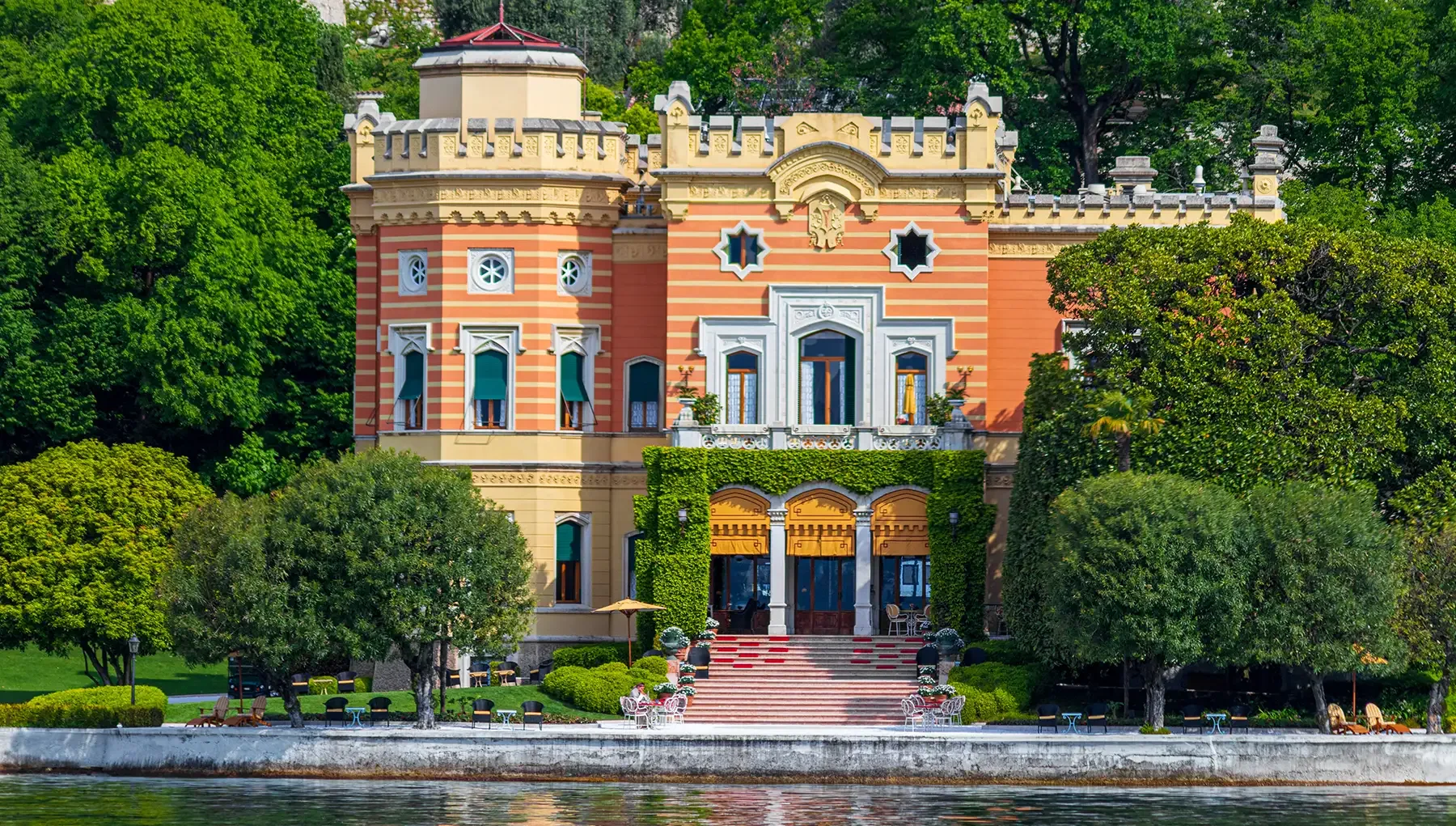 Historic lakeside villa with striped orange facade, lush gardens and waterfront steps.