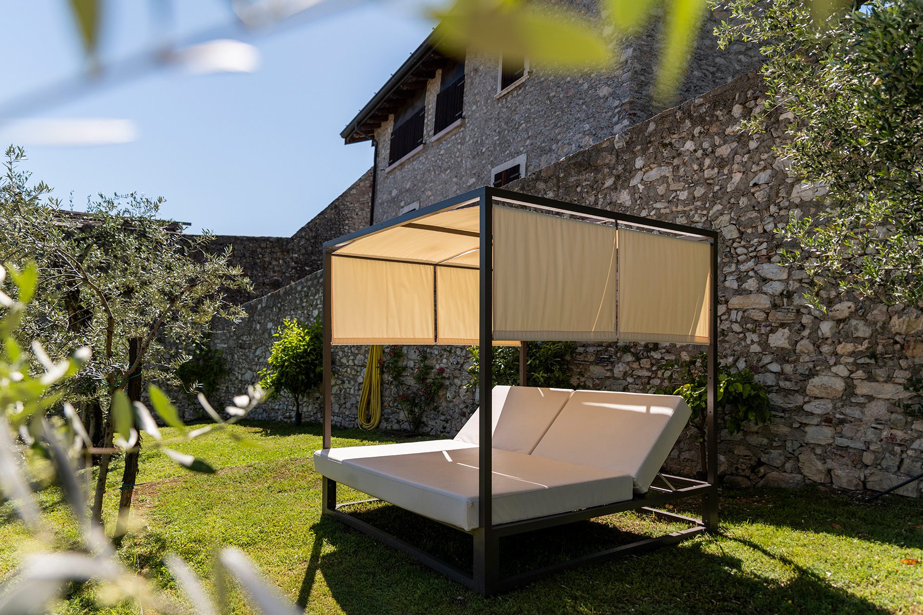 Letto a baldacchino da esterno in giardino con ulivi e muro in pietra
