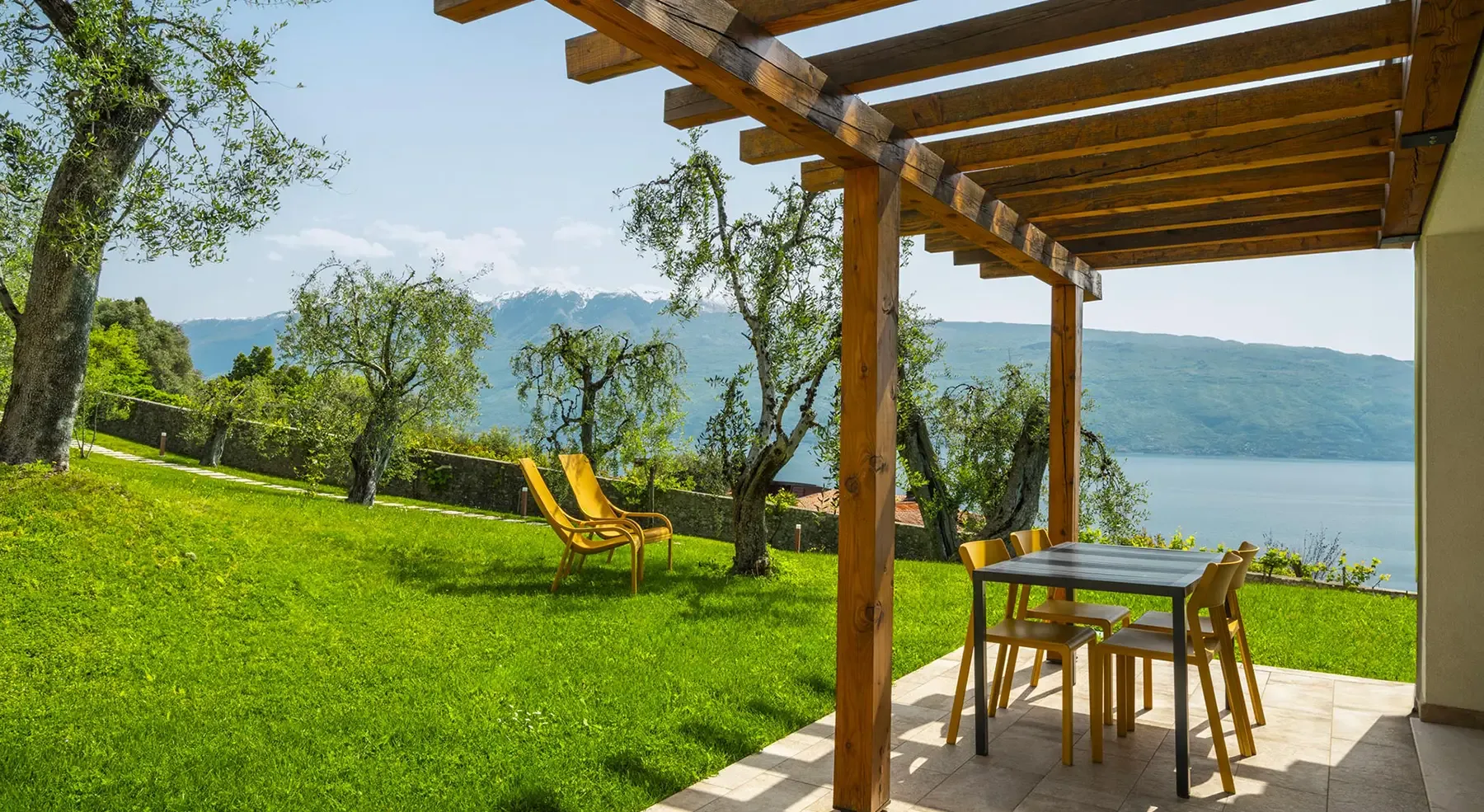 Terrazza con pergolato in legno, tavolo e vista lago tra ulivi e montagne.